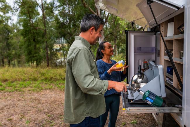 A couple prepares food at a campervan's slide-out outdoor kitchen in a grassy forest clearing.