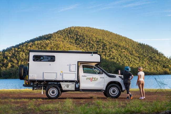 Two people stand next to a white 4WD campervan parked by a lake, with a forested hill in the background.