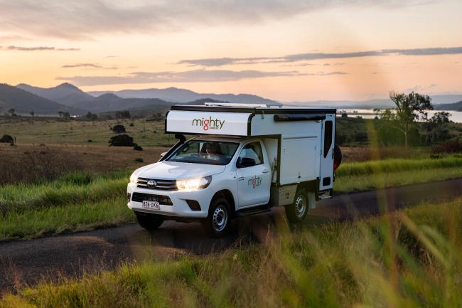 White Mighty campervan driving on a rural road at sunset with hills in the background.