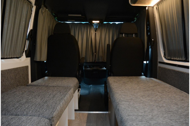 Campervan interior with grey fabric bench beds, a central aisle, black front seats, and matching grey privacy curtains.