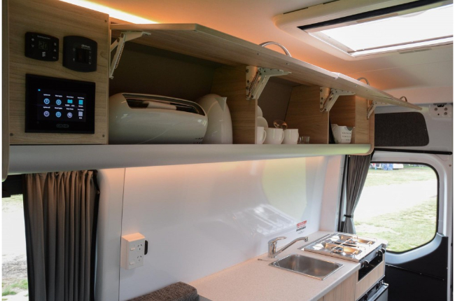 Campervan kitchen with open overhead cabinets, a digital control panel, a stainless steel sink, and a cooktop.