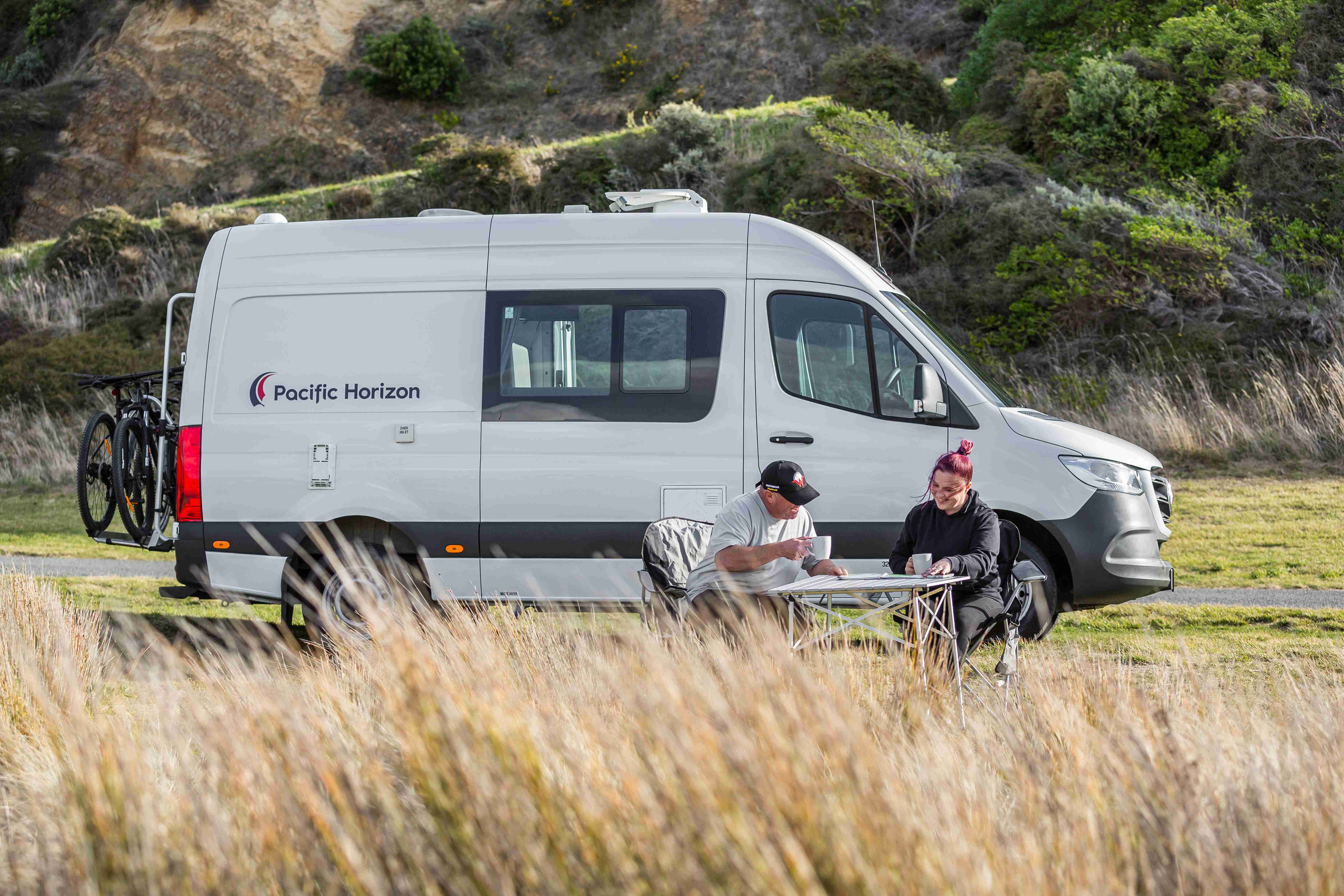Two people sitting at a folding table outside a white Pacific Horizon campervan parked in a grassy field.