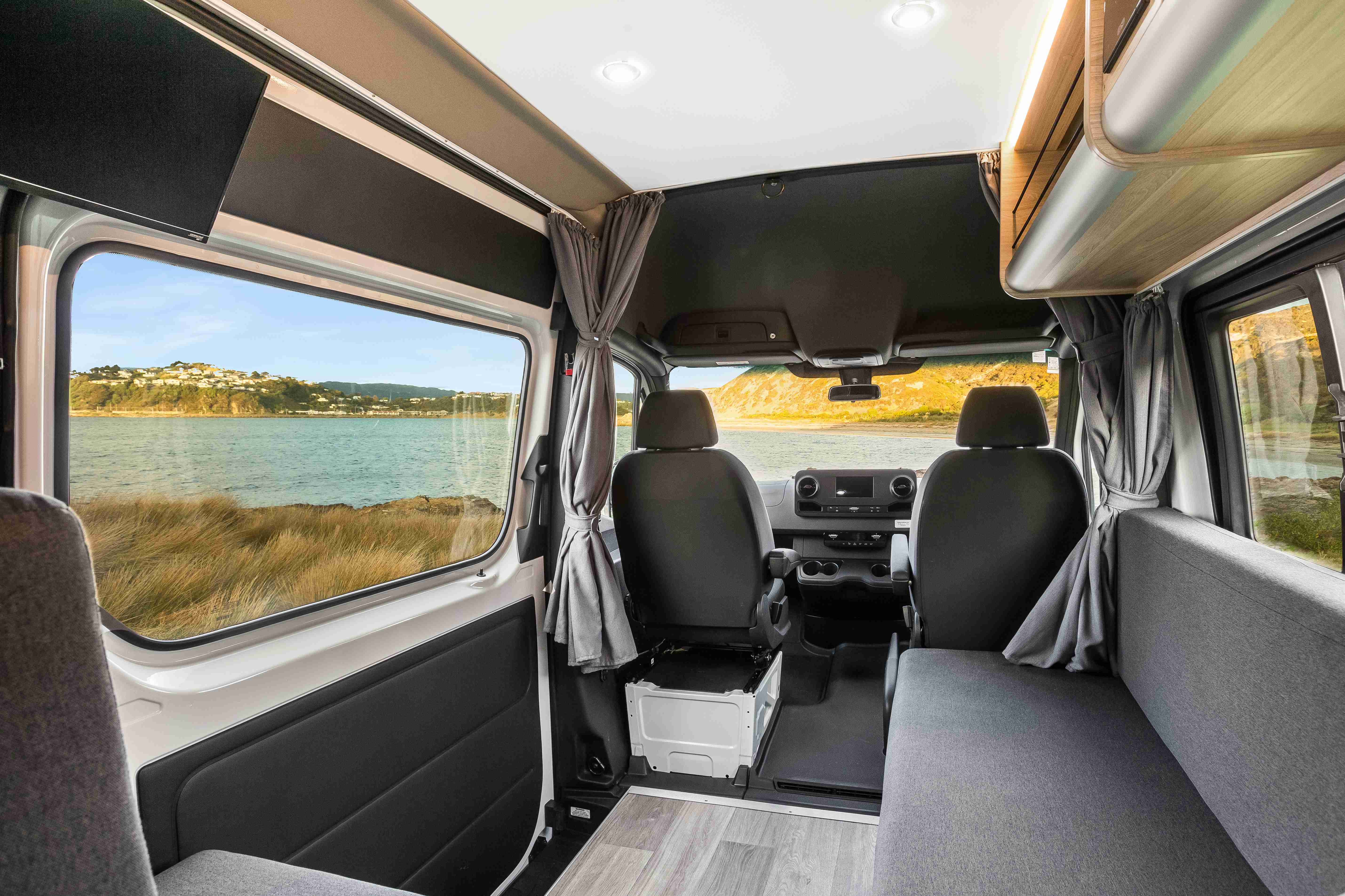 Interior of a campervan looking forward, with grey seats and a large window overlooking a coastal landscape.