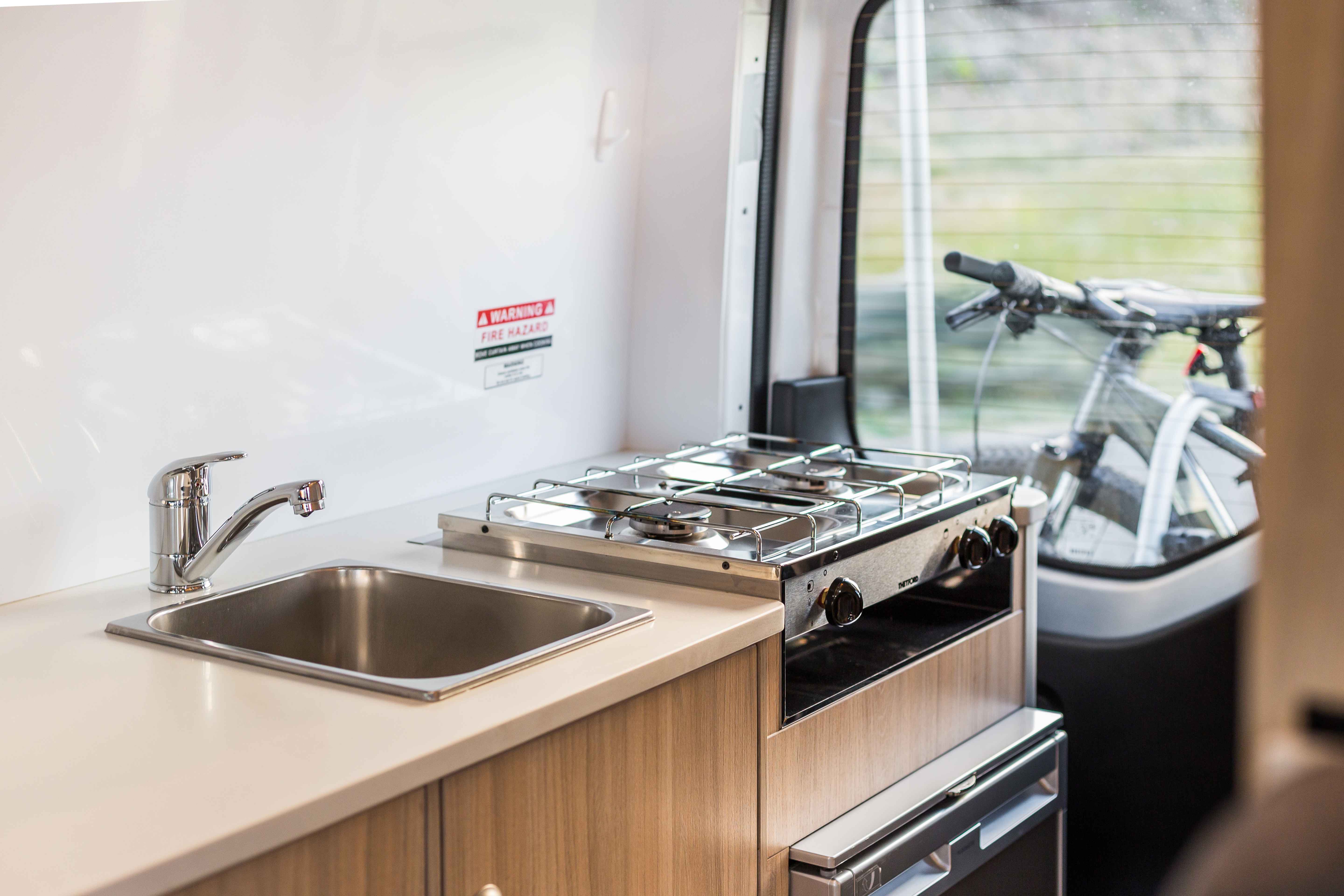 Compact campervan kitchen with a stainless steel sink, two-burner gas stovetop, and light timber cabinets.