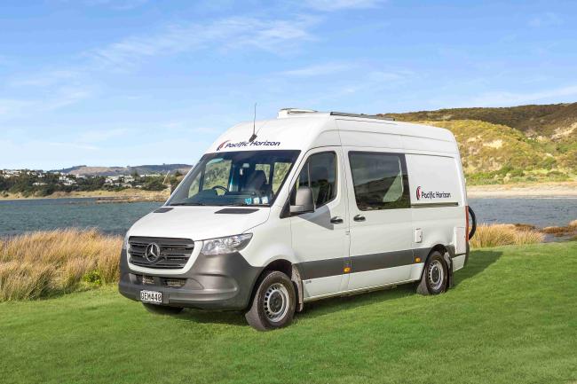 White Mercedes campervan with Pacific Horizon branding parked on a grassy field next to a coastal bay.