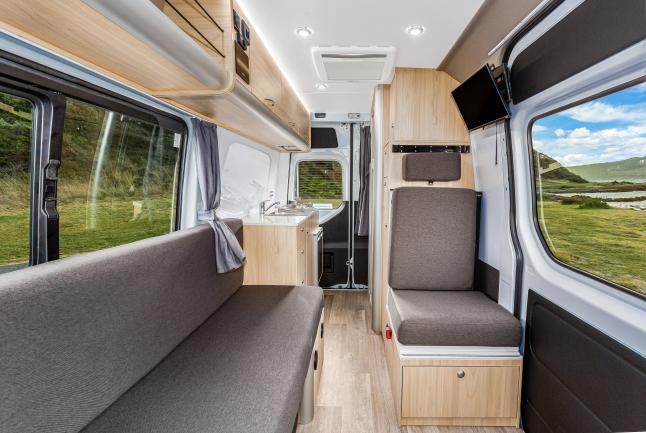 Modern campervan interior featuring grey upholstered seats, light timber cabinets, and windows showing outdoor greenery.
