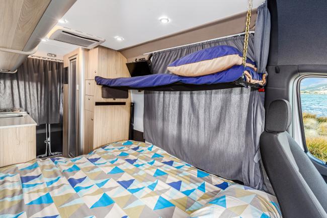 Campervan interior featuring a double bed with a geometric quilt and a suspended single bunk above.