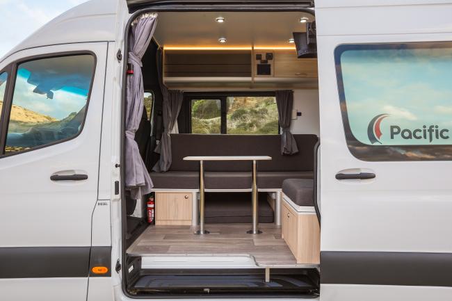 Campervan interior with grey bench seats, a central dining table, and light timber-look flooring and cabinets.