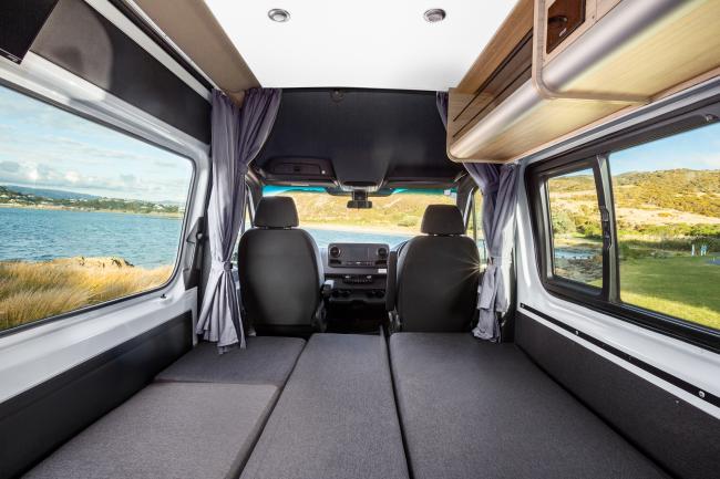 Campervan interior looking forward, featuring grey bed platforms, front seats, and side windows with coastal views.