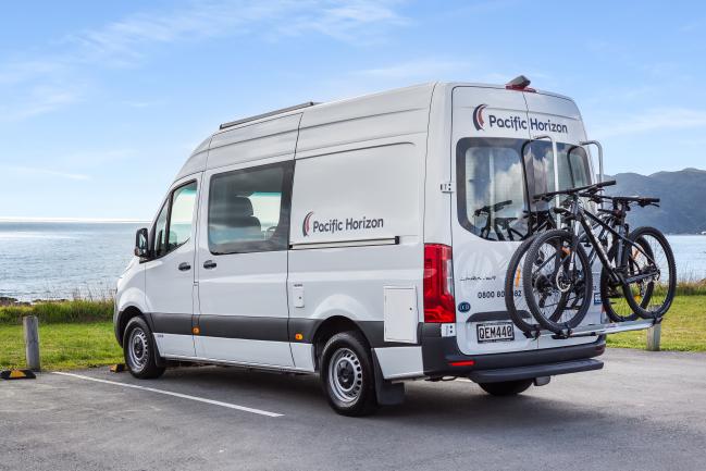 White Pacific Horizon campervan with two mountain bikes on a rear rack, parked in a coastal car park.