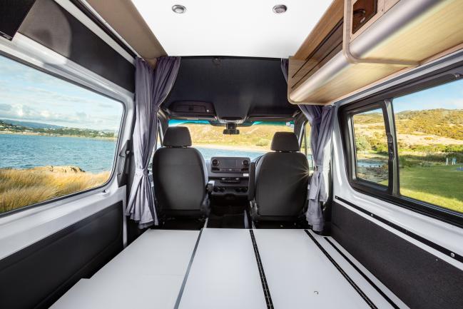 Campervan interior showing a flat white bed platform, two front seats, and coastal views through the windows.
