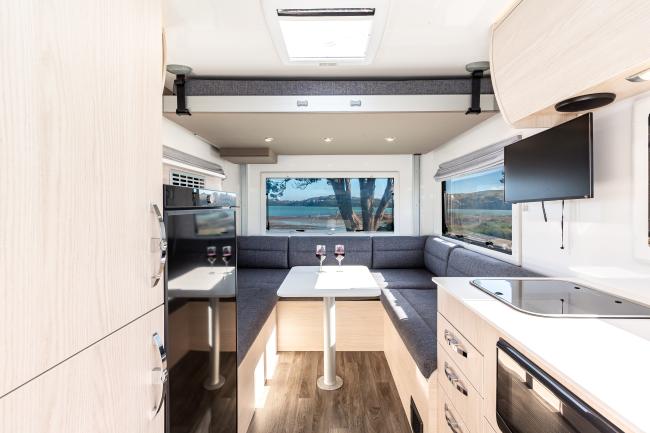 Modern campervan interior with light timber cabinetry, grey U-shaped dining area, kitchenette, and scenic window view.