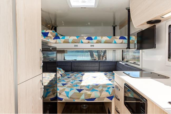 Campervan interior featuring double bunk beds with blue geometric linen, light cabinetry, and a compact kitchenette.