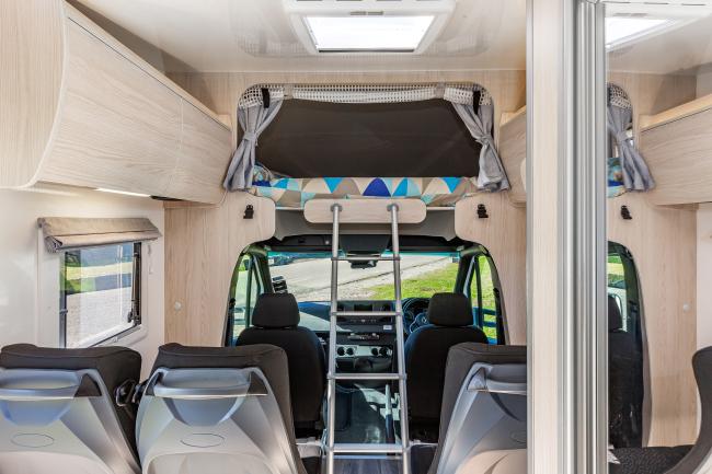 Campervan interior showing passenger seats, a ladder to an over-cab bed, and light timber cabinetry.