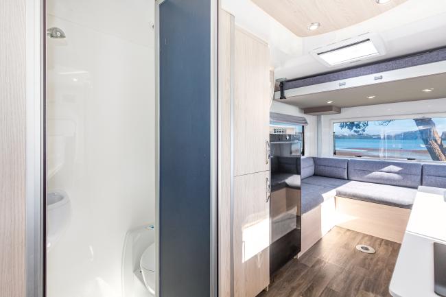 Campervan interior showing a bathroom on the left, timber-look cabinetry, and a grey U-shaped lounge in the rear.