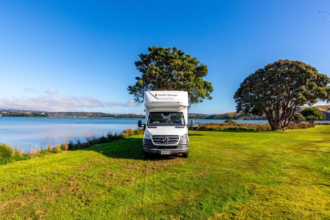 White campervan parked on a grassy field next to calm water under a blue sky with trees.