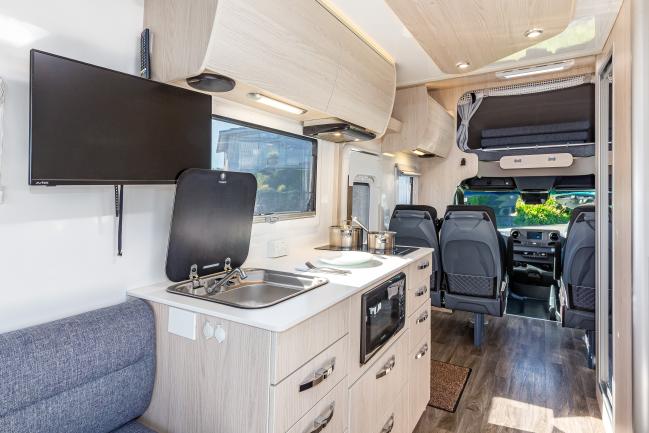 Modern campervan interior featuring a compact kitchenette with sink and microwave, wall-mounted TV, and cab seats.