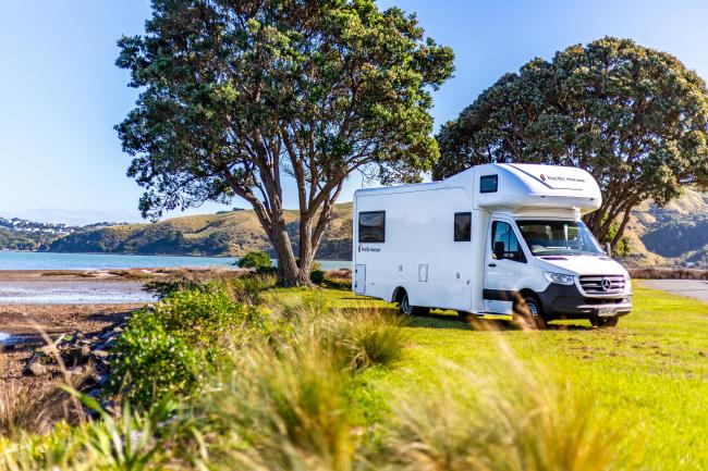 White Mercedes-Benz campervan parked on a grassy bank next to a coastal inlet under large leafy trees.
