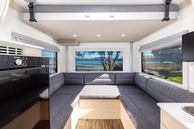 Campervan interior featuring a grey U-shaped lounge, a drop-down bed overhead, and windows overlooking water.