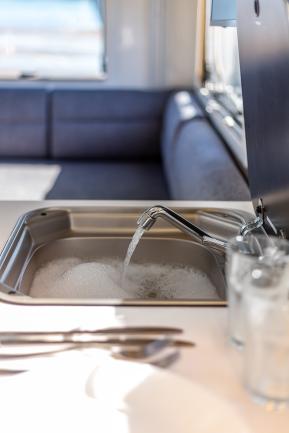 Water running from a chrome tap into a soapy stainless steel sink inside a campervan with seating in the background.
