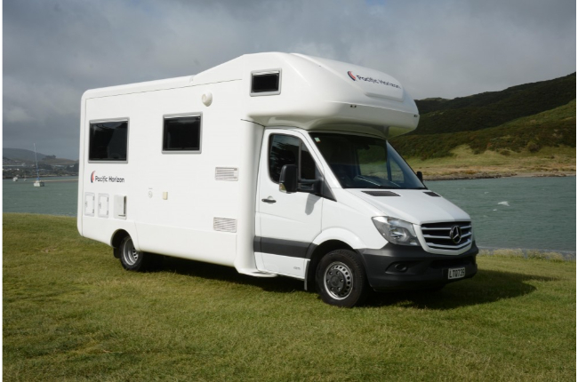 White Mercedes-Benz campervan parked on a grassy bank next to water with hills in the background.