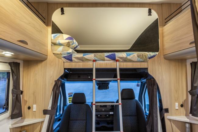 Campervan interior showing an over-cab loft bed with a ladder above the driver and passenger seats.
