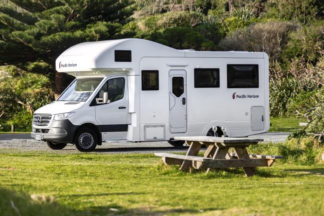 White Pacific Horizon campervan parked beside a grassy reserve with a timber picnic table and dense trees behind.