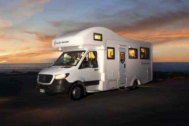 White Pacific Horizon campervan parked by the ocean at sunset with interior and exterior lights illuminated.