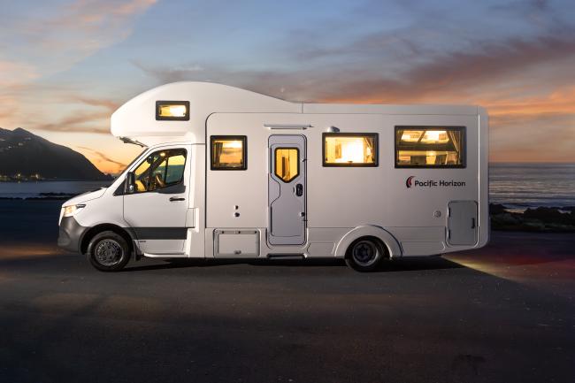 Side view of a white Pacific Horizon campervan parked near the coast at dusk with glowing interior lights.