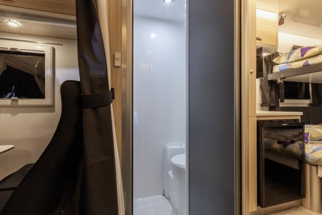 Campervan interior showing a compact bathroom flanked by a black seat and bunk beds with a built-in fridge.