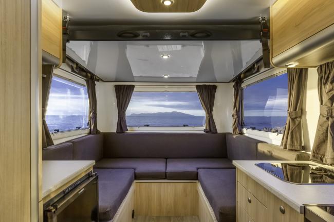 Campervan interior showing a U-shaped lounge with grey cushions, light timber cabinets, and windows with ocean views.