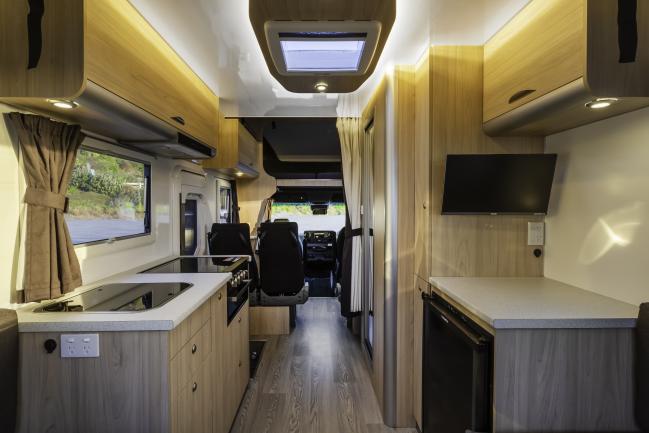 Interior of a modern campervan with light timber-look cabinetry, kitchen facilities, TV, and aisle leading to the cabin.