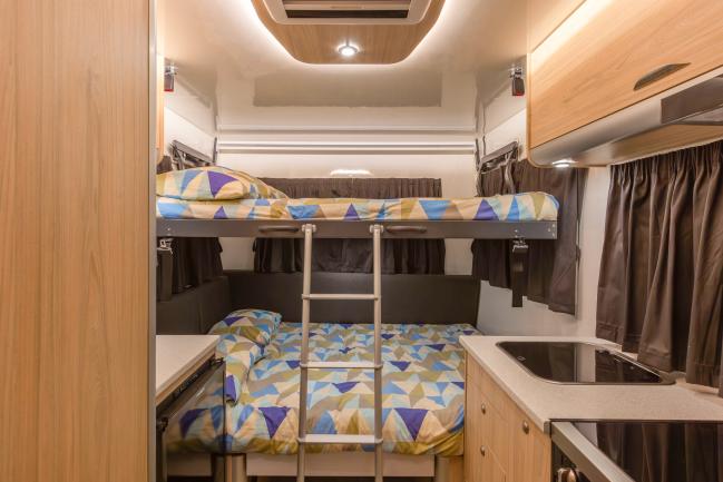 Campervan interior with timber cabinets, a kitchenette, and bunk beds with colourful geometric bedding and a ladder.