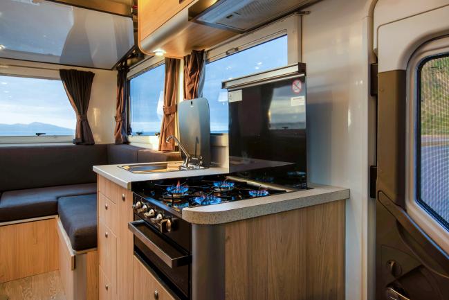 Campervan interior showing a kitchen with lit gas burners, timber cabinets, and a grey lounge area in the background.