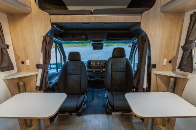 Campervan interior showing swivelled cabin seats facing two small tables, timber-look panelling, and an over-cab bed.