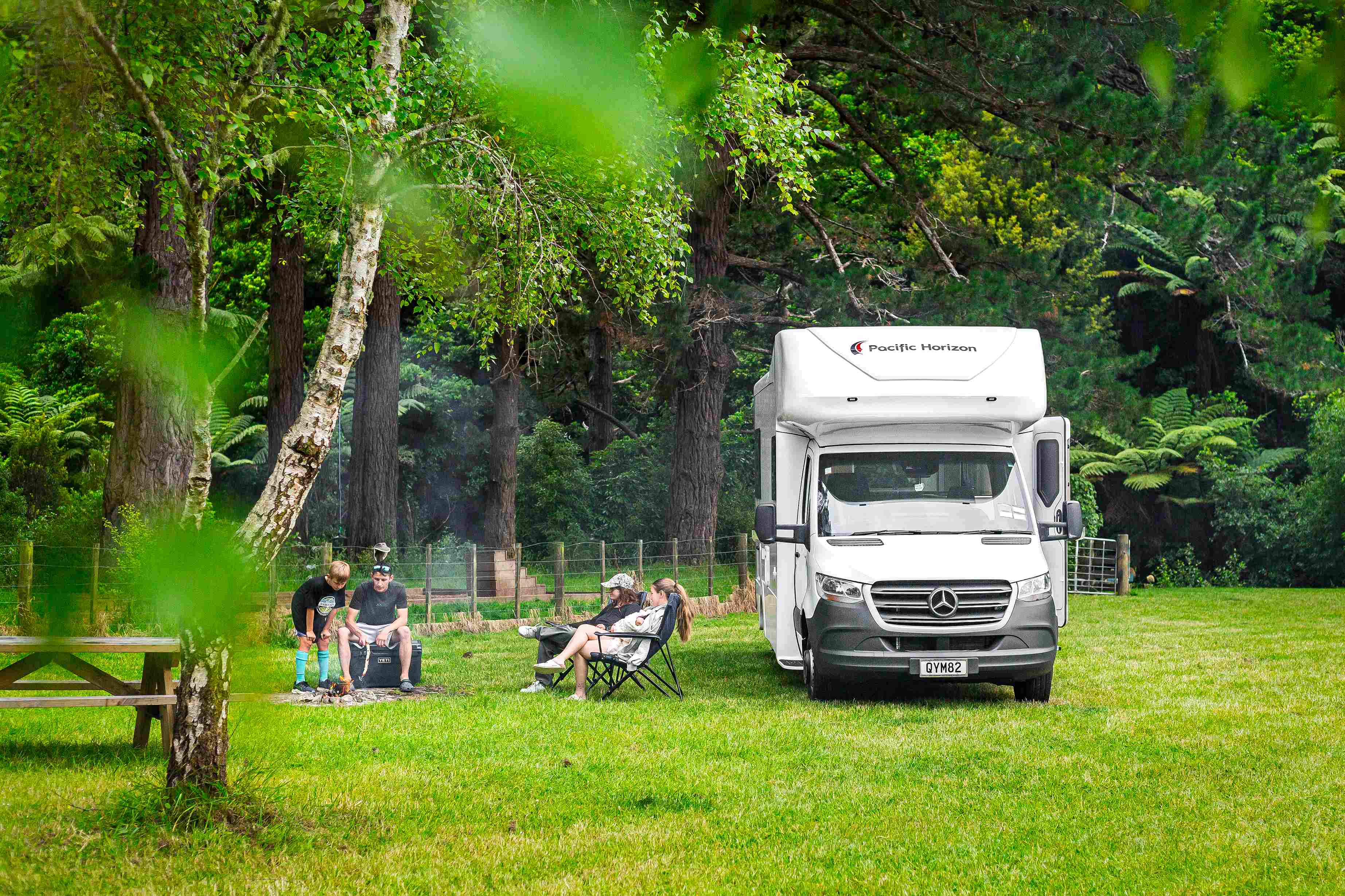 Family by a campfire in a grassy clearing next to a white campervan parked near a forest.