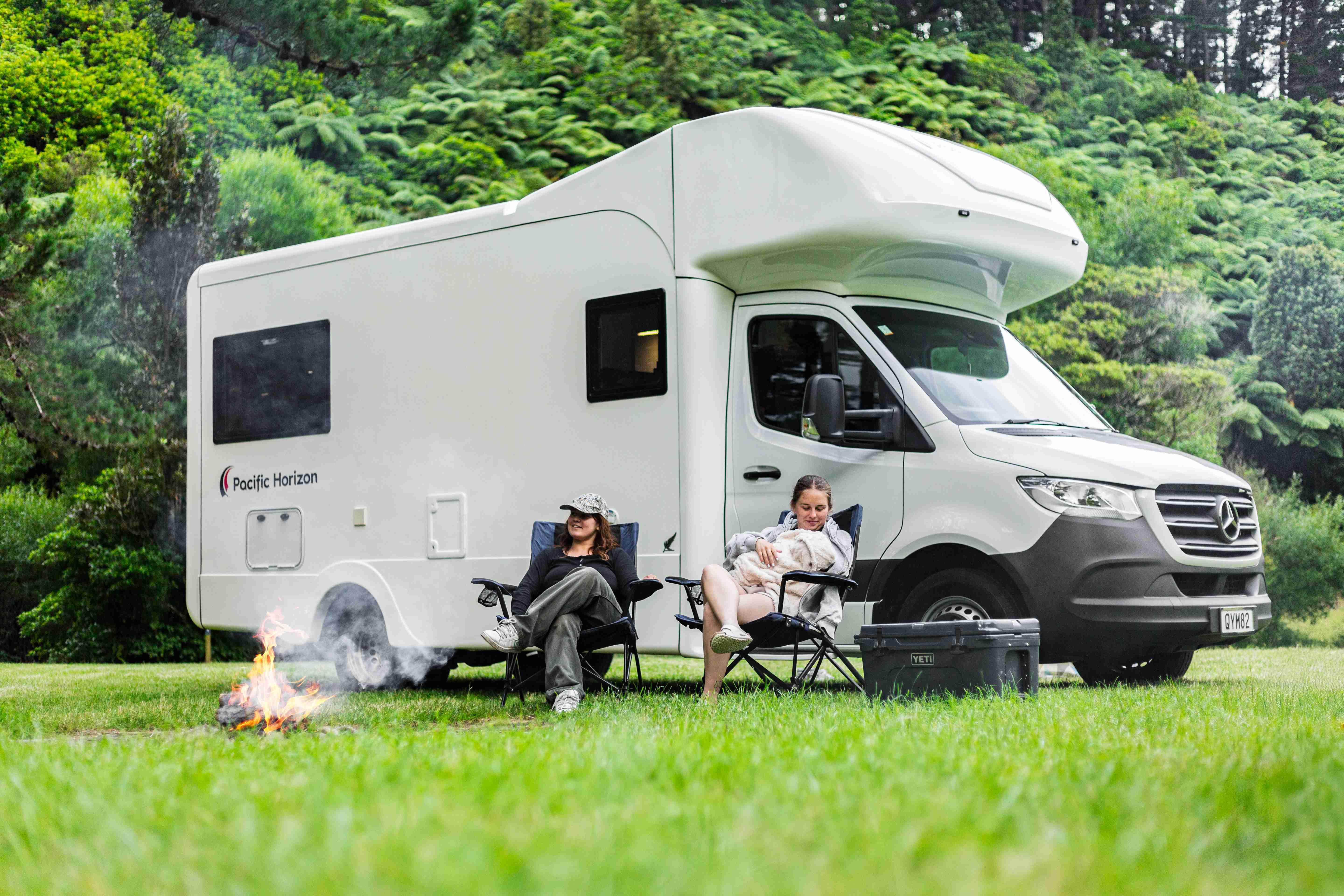 Two people relax by a campfire next to a white campervan parked in a grassy forest clearing.
