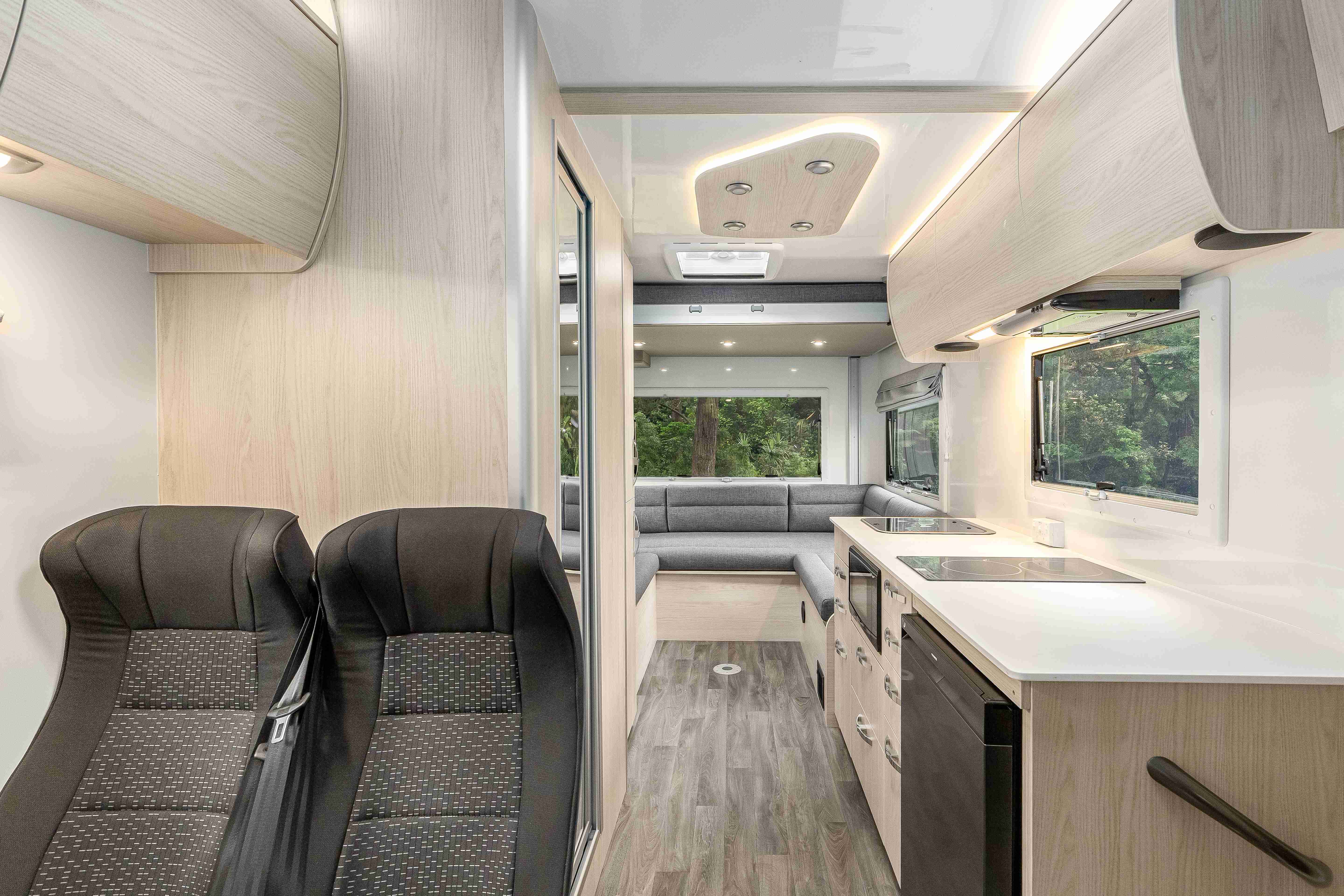 Modern campervan interior with dark grey front seats, a light-coloured kitchenette, and a rear grey fabric lounge.