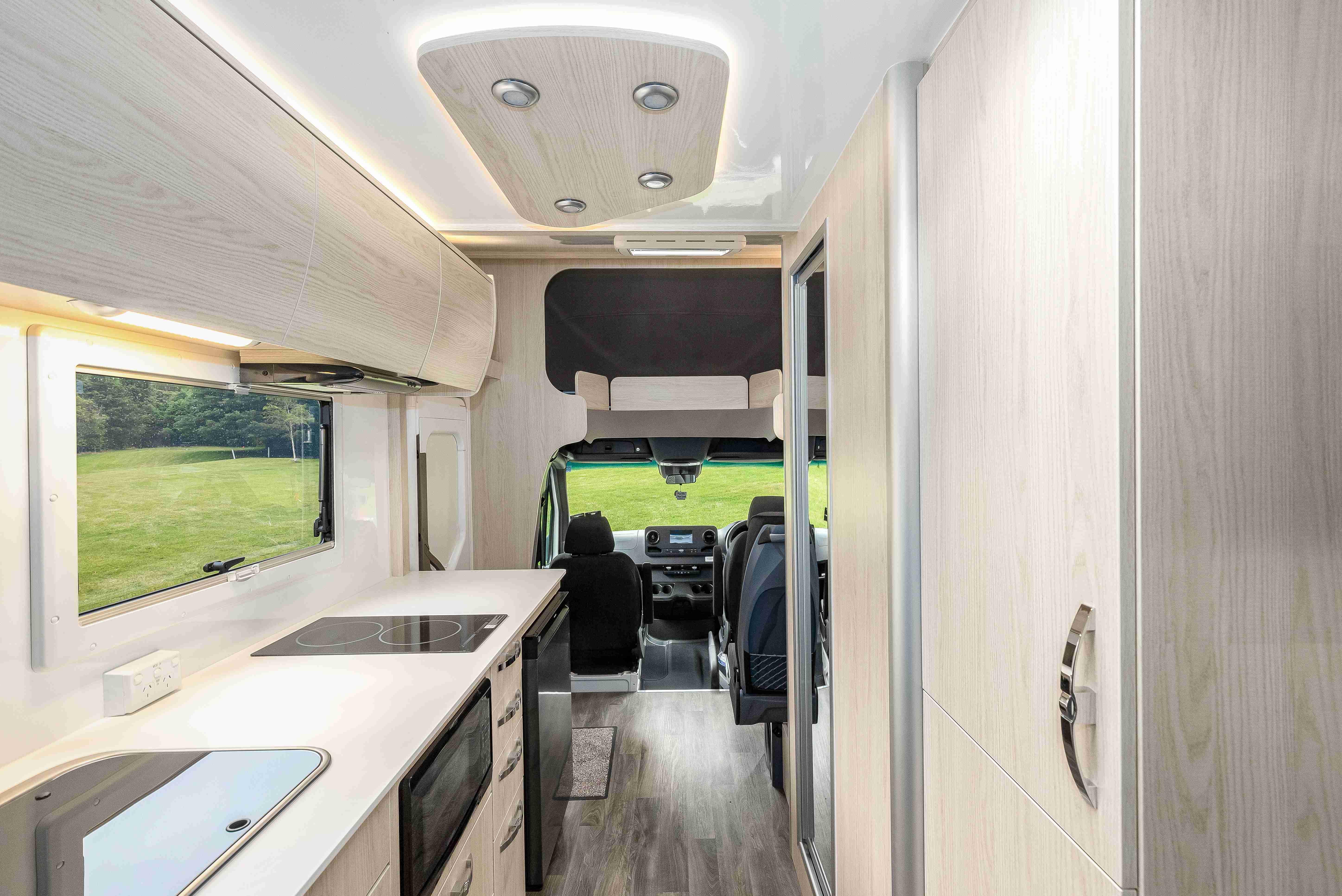 Modern campervan interior featuring light timber-grain cabinets, a kitchen benchtop, and view of the front driver cabin.