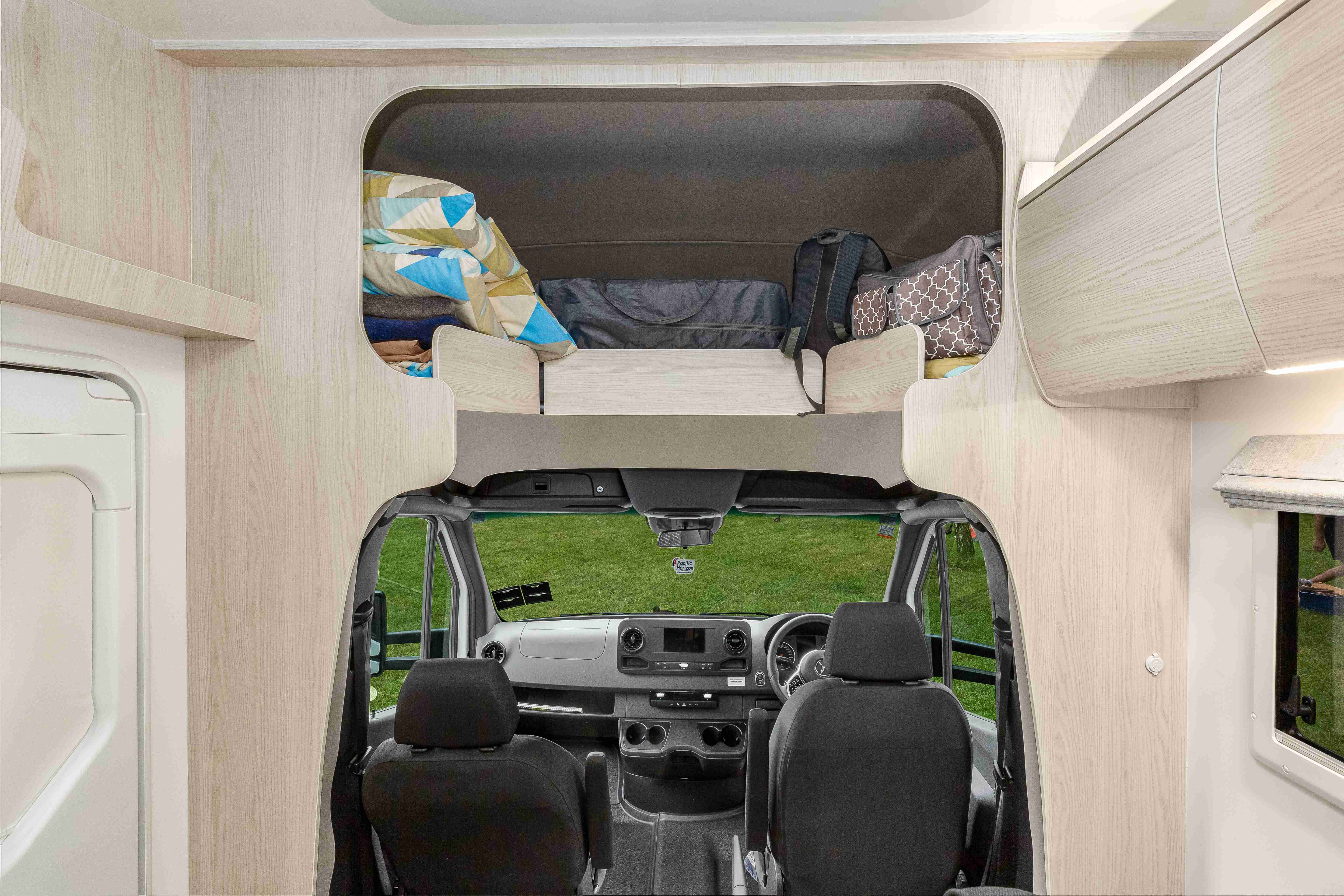 Campervan interior looking forward to the front seats, with an over-cab storage compartment holding luggage and bedding.