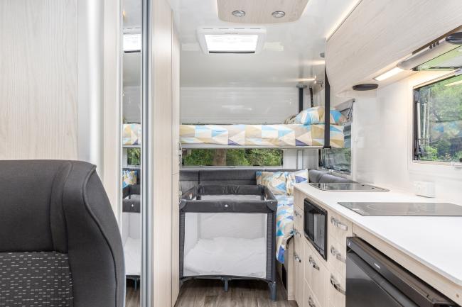 Campervan interior featuring a kitchenette, a grey travel cot, and an overhead bunk bed.