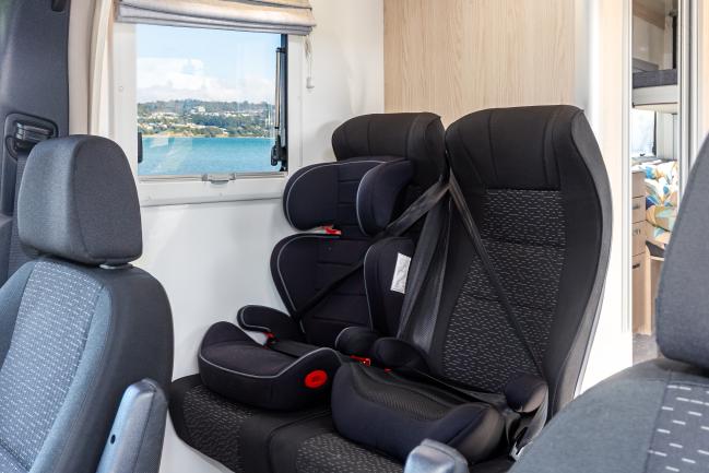 Two black child safety seats secured on passenger seats inside a campervan, next to a window with a coastal view.