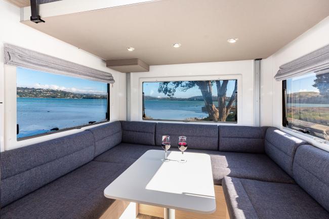 Campervan interior with grey U-shaped seating, a white table with two wine glasses, and windows overlooking a bay.