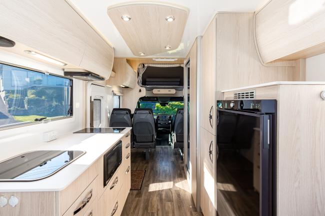 Modern campervan interior with light timber-look cabinets, kitchen counter with sink, black fridge, and cabin seats.