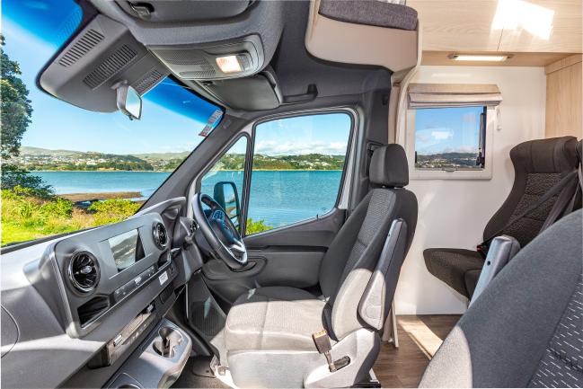 Campervan cabin interior with grey seats, showing a scenic coastal view through the front and side windows.