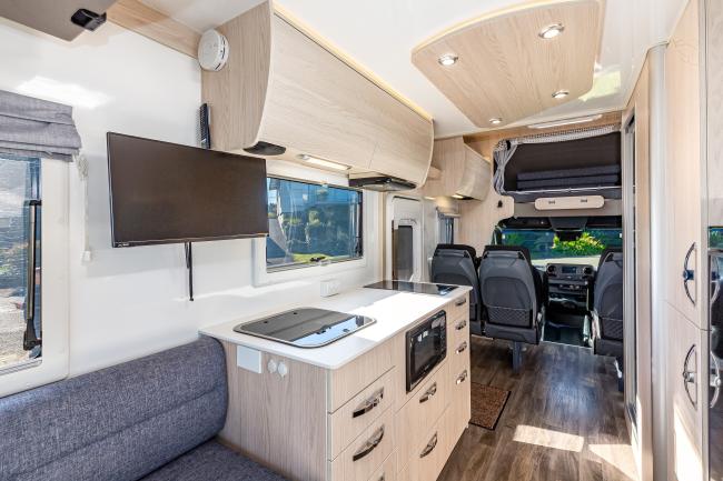Campervan interior featuring light timber cabinetry, a kitchenette with microwave, wall-mounted TV, and grey seating.