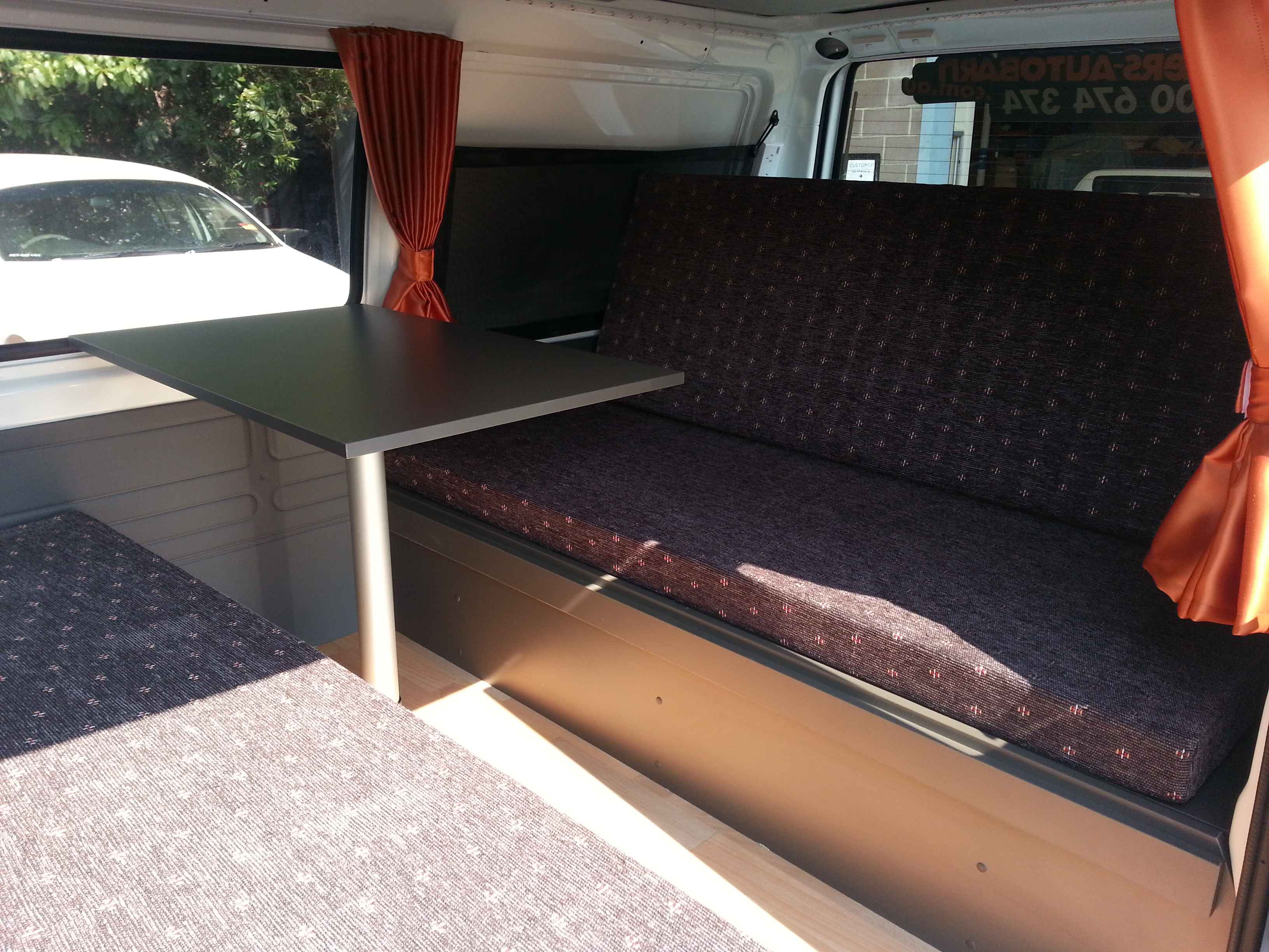 Campervan interior featuring a black pedestal table between two dark patterned fabric bench seats.