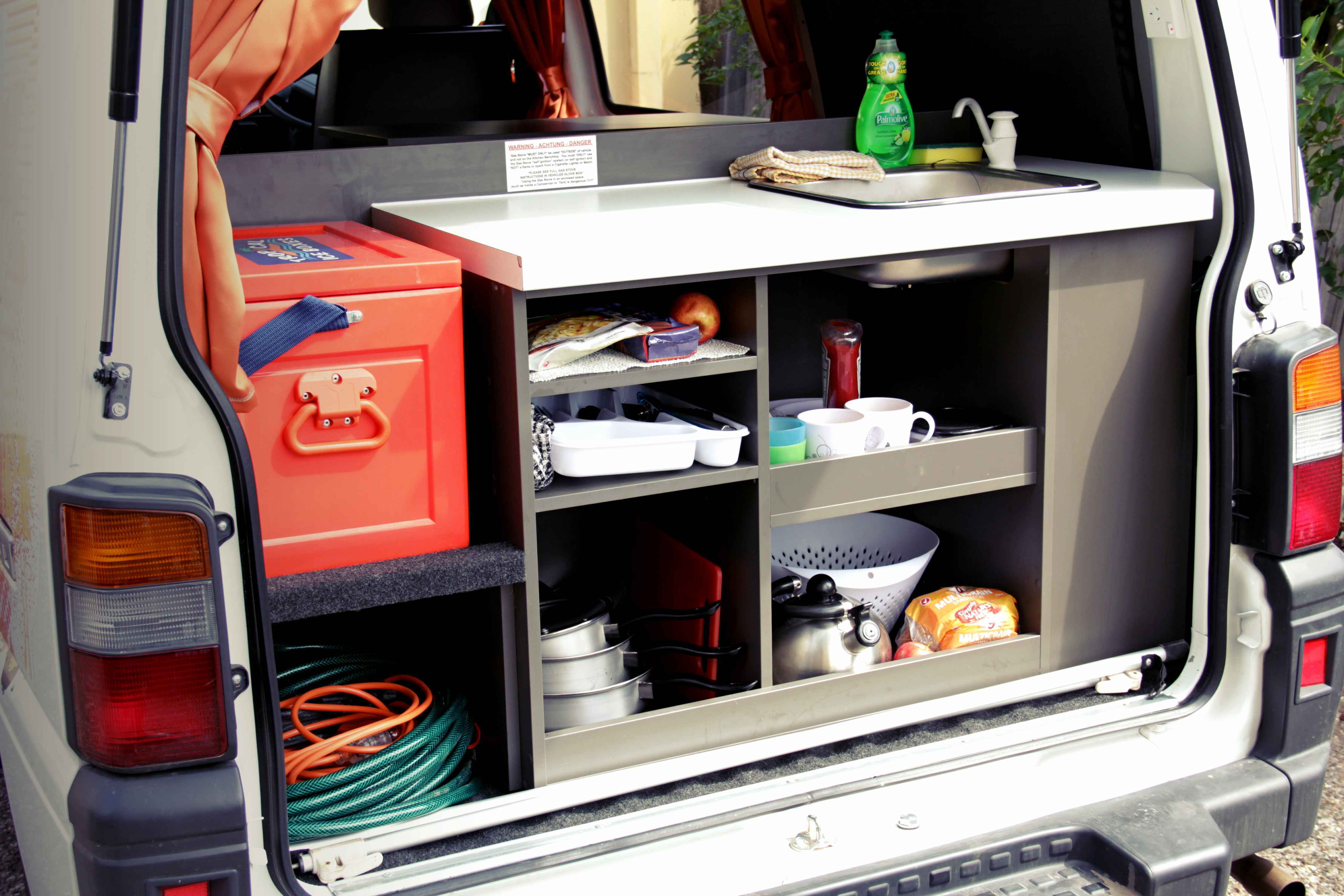 Campervan rear kitchen setup with a built-in sink, red cooler box, storage shelves, and cooking equipment.