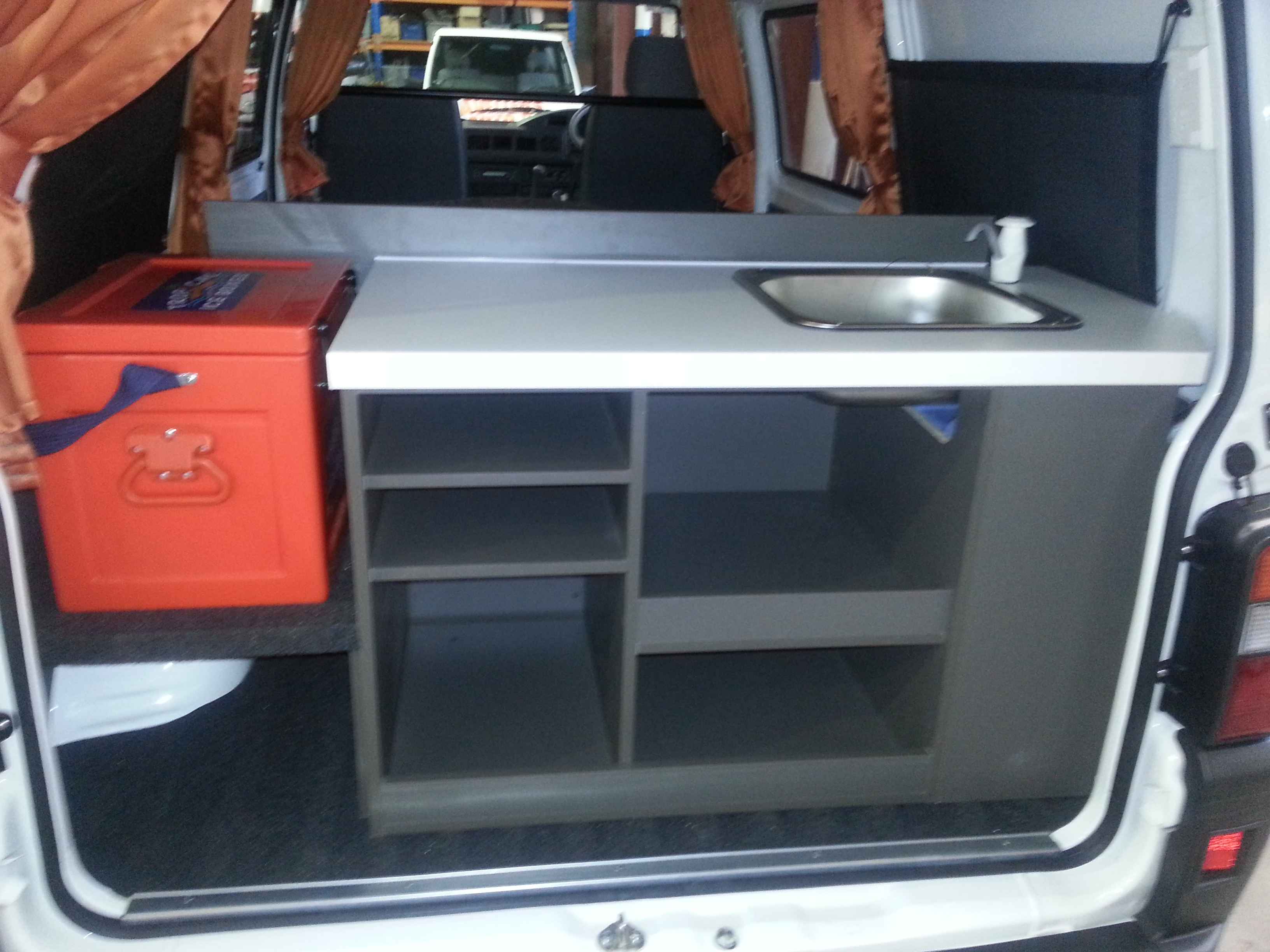 Campervan rear interior with grey cabinets, a white benchtop, small sink, and an orange cooler box.