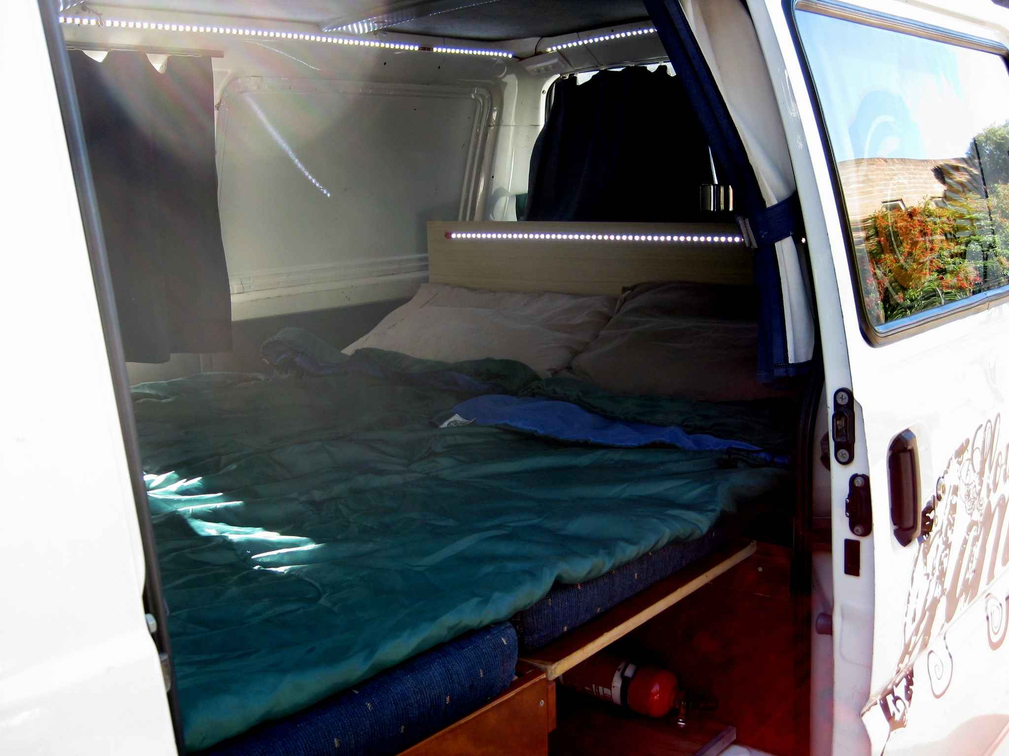 Inside a white campervan featuring a bed with a green sleeping bag, pillows, and LED strip lighting.